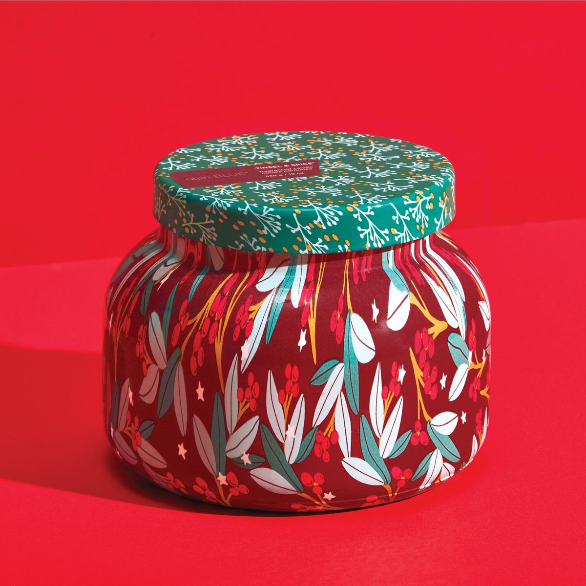Capri Blue Tinsel & Spice Winter Bazaar Candle, festive red jar with leaves, berries, and stars, green lid, on red background.