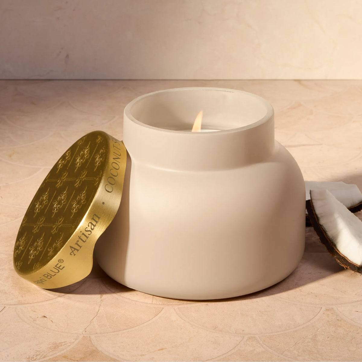 Capri Blue Coconut Santal Terrazza Velluto Candle, lit, with gold lid & coconut pieces on a beige surface