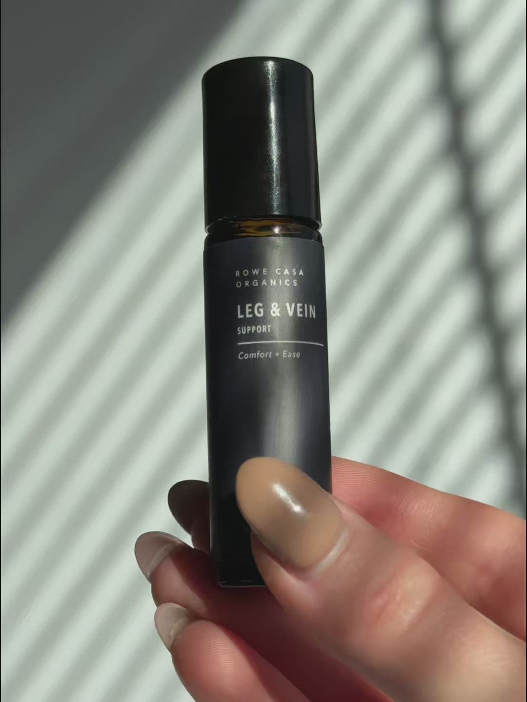 Hand holds a dark roller bottle of Rowe Casa Organics Leg & Vein Support 10ml. Label reads 'Comfort + Ease'.